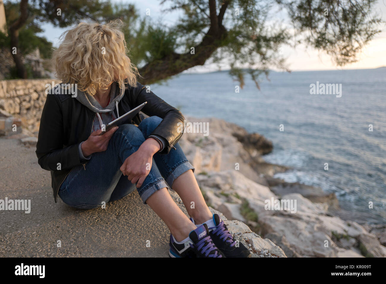 Bella ragazza bionda con capelli ricci di posti a sedere su pietre in  prossimità del mare con la compressa in mani e guardando in lontananza Foto  stock - Alamy