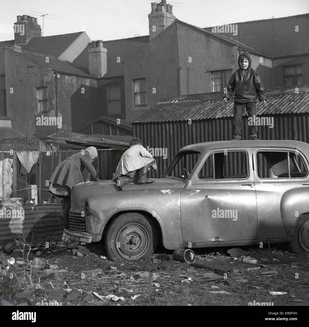 Anni sessanta, parco giochi urbano, scruffy giovani bambini giocano su una vecchia auto abbandonate su all interno della città wasteland sul retro del case a schiera, Inghilterra. Foto Stock