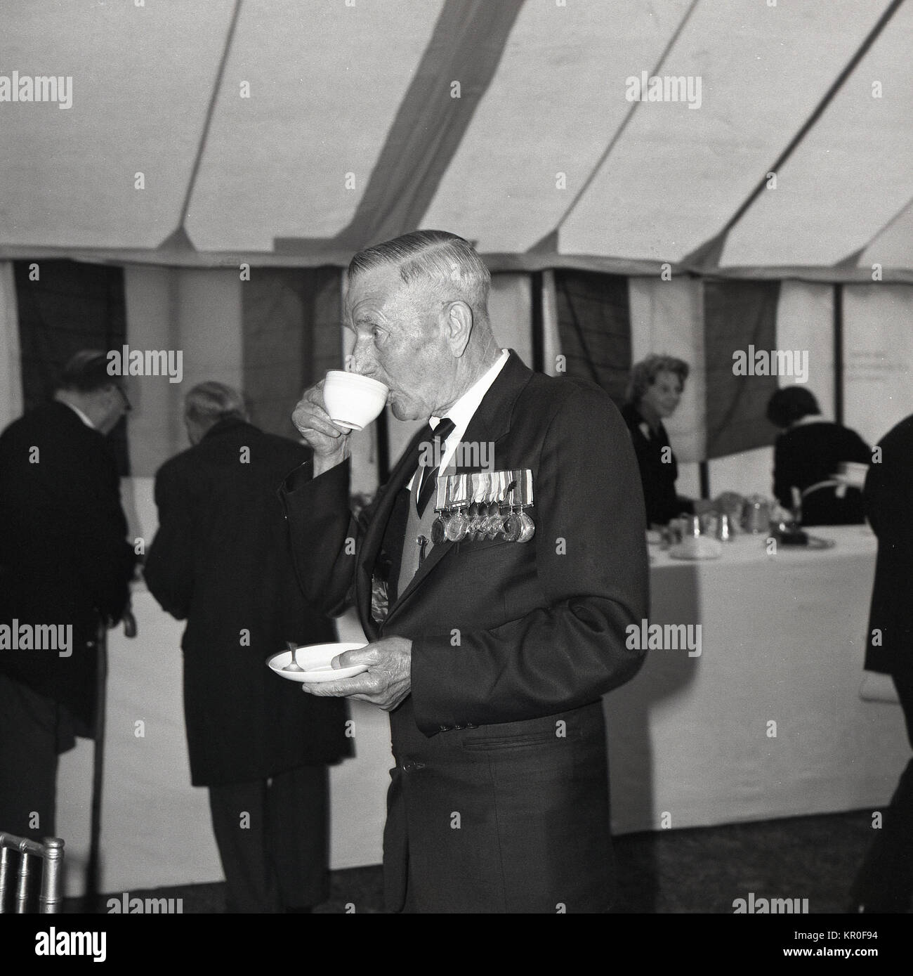Anni sessanta, Inghilterra, British WW2 veterano militare con medaglie gustando una tazza di tè in una ri-unione evento in un rettangolo di selezione. Foto Stock
