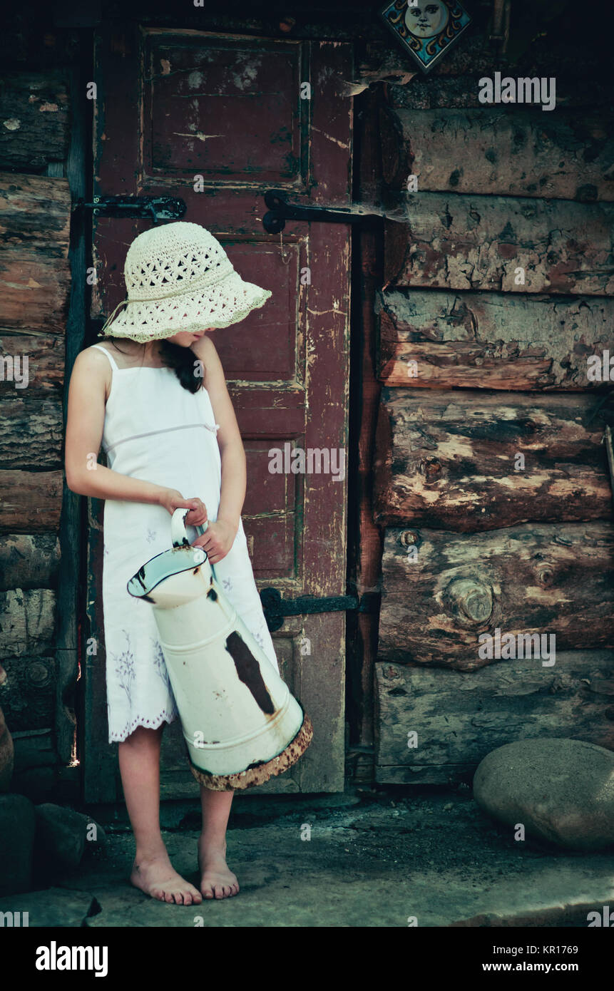 Ragazza giovane 5-7 anni tenendo una brocca e in piedi di fronte ad una vecchia casa in legno. Retrò e vintage in cerca dell'immagine. Foto Stock