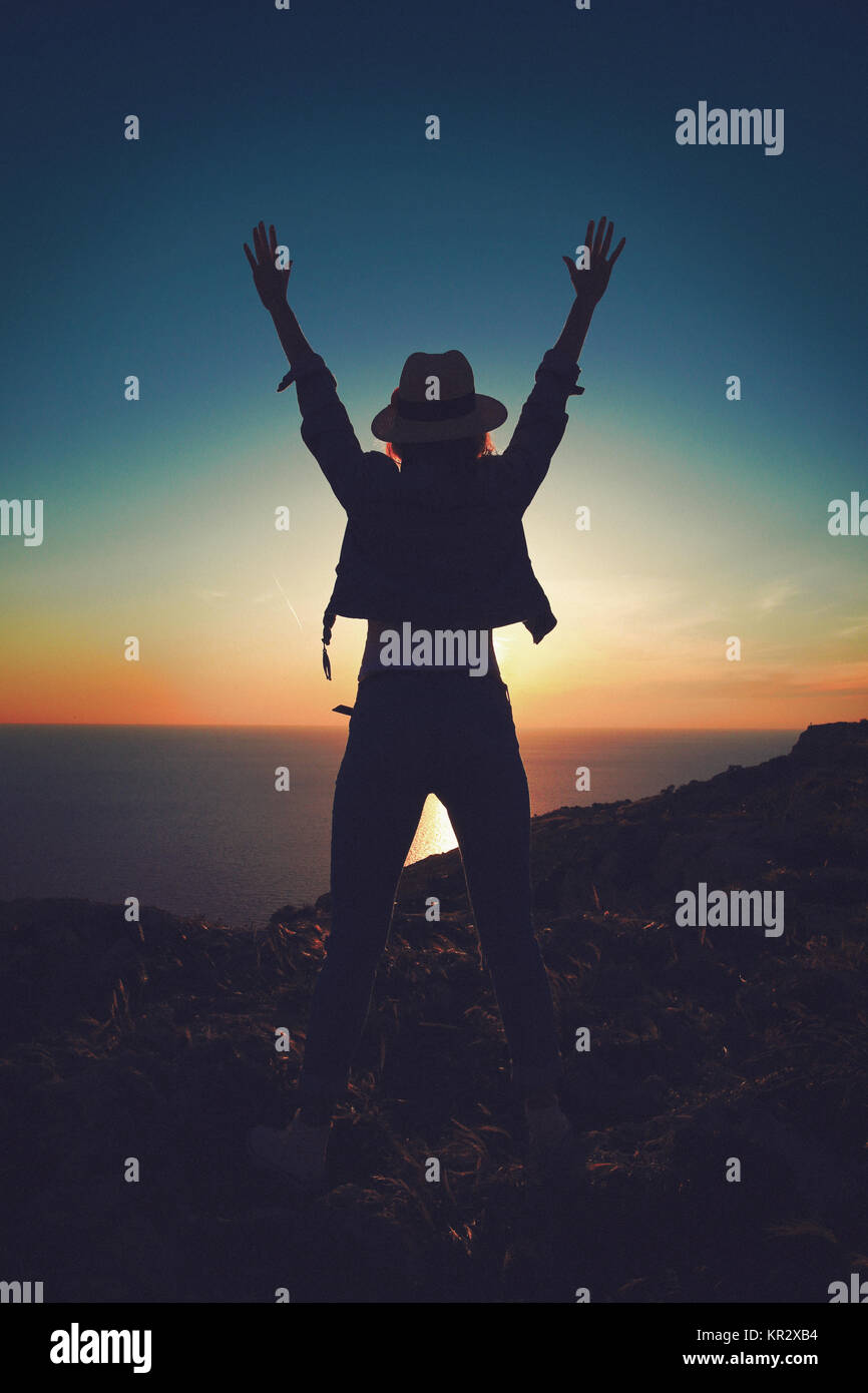 Carefree woman in hat silhouette in alto le mani nel tramonto sulla scogliera sul mare Foto Stock