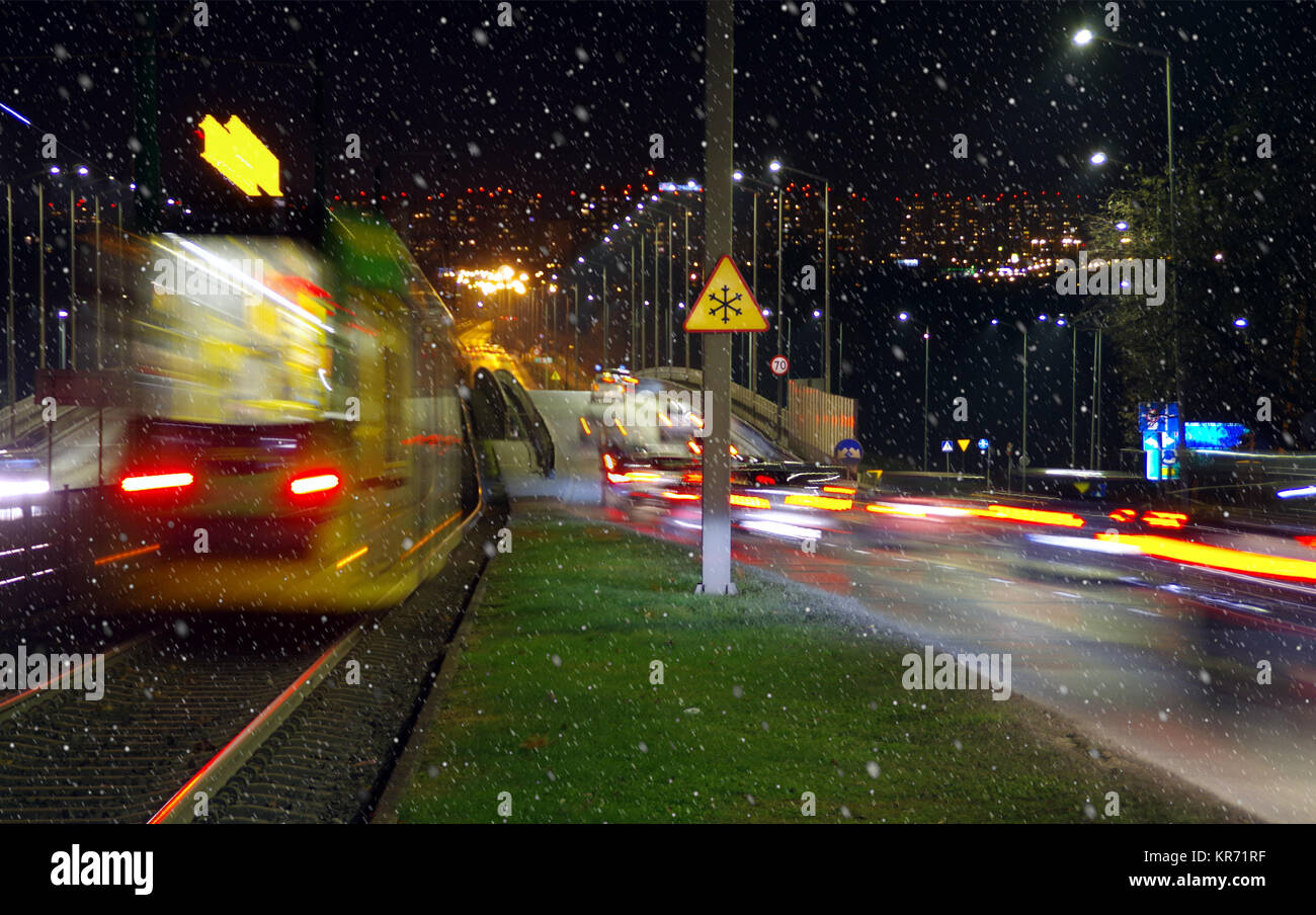 Prima nevicata in città di notte. Luci sfocate automobili e tranviaria cittadina sulla strada con i fiocchi di neve in primo piano. Foto Stock