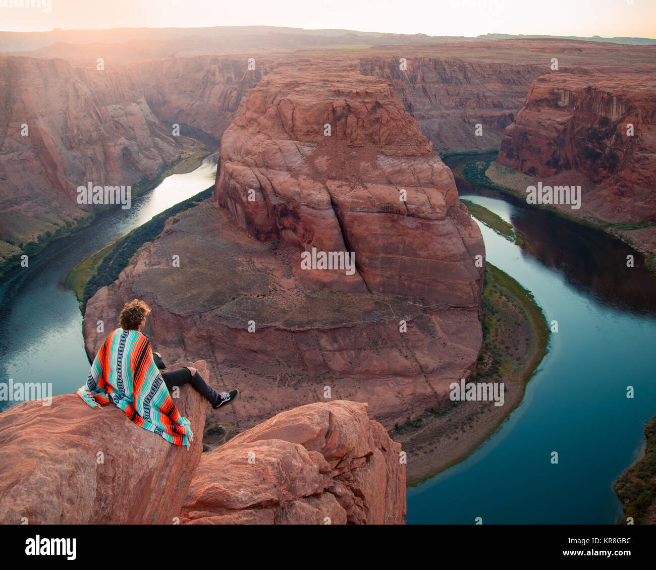 Giovane uomo seduto sulla sporgenza di roccia guardando oltre la curva a ferro di cavallo in Arizona, Stati Uniti durante il tramonto Foto Stock