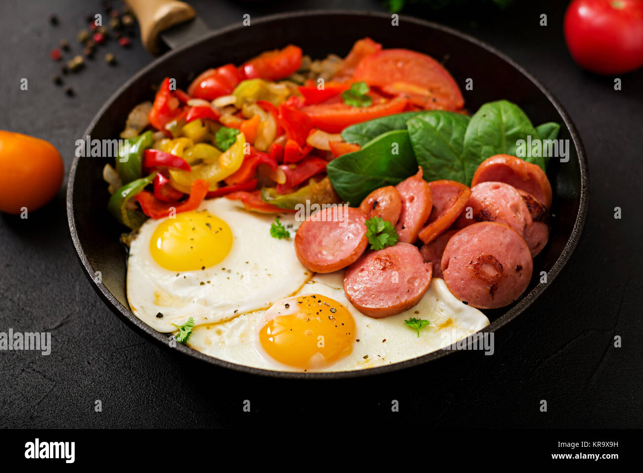 La prima colazione. Uova fritte con salsiccia e verdure in una padella su uno sfondo nero in stile rustico. Foto Stock