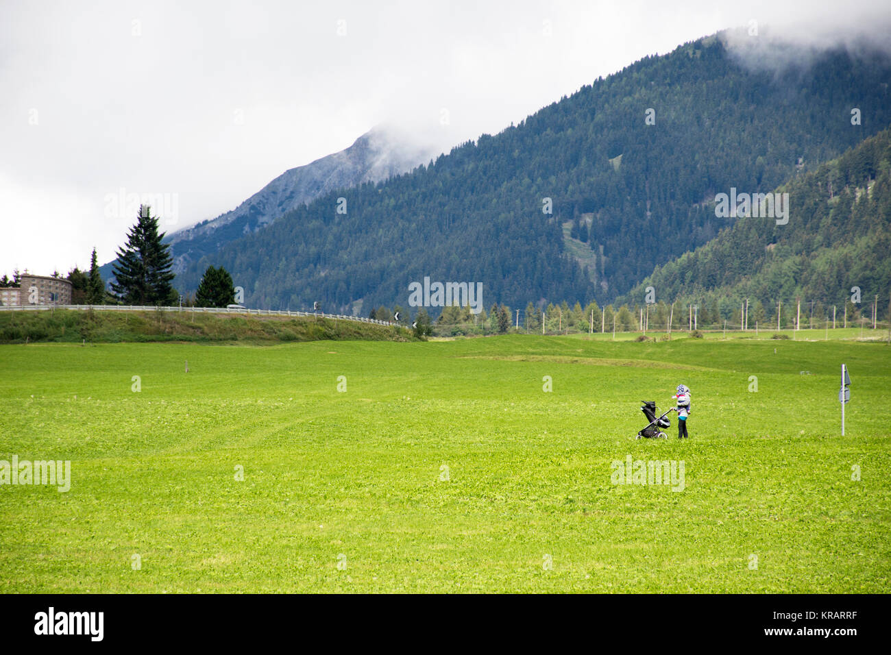 Madre italiana di spingere un passeggino nel campo di erba accanto alla strada vicino a Hyde park in Burgusio Malles Venosta a Malles Venosta, in val Venosta, in Trentino-Alt Foto Stock