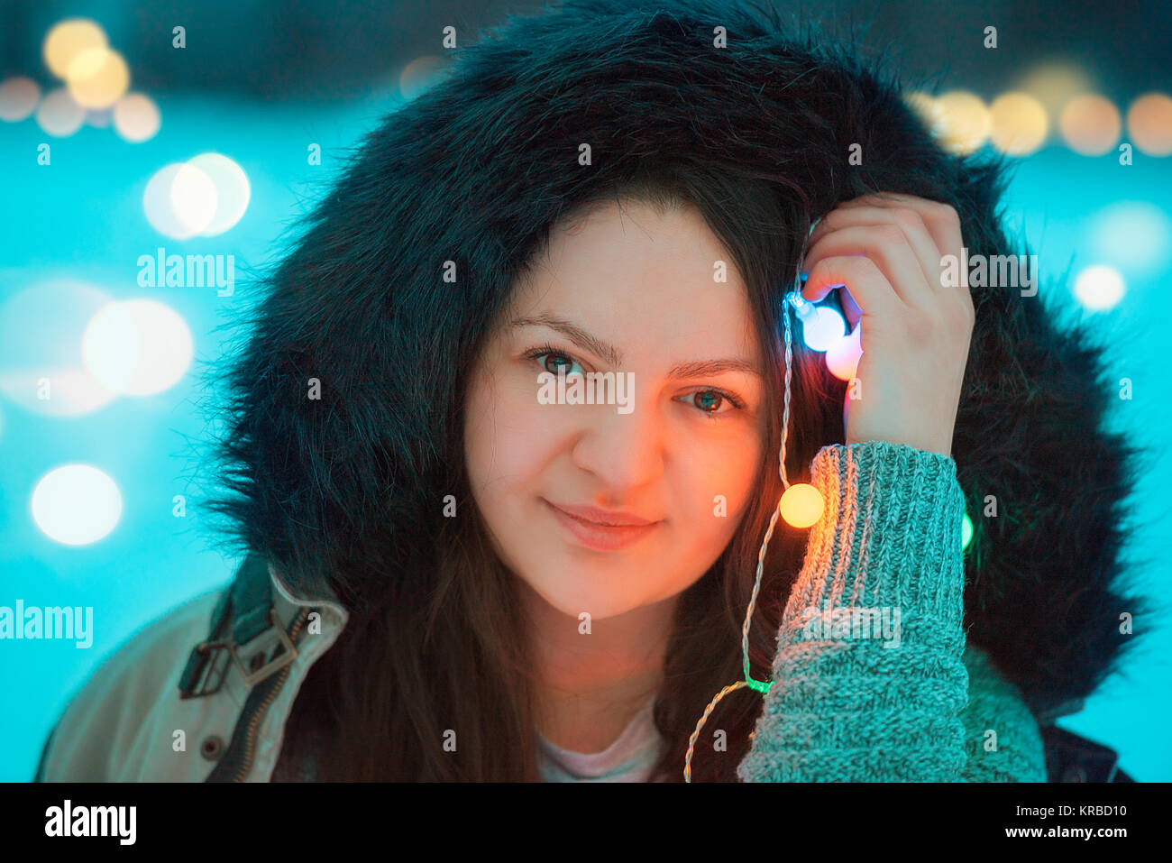 Ritratto di un giovane sorridente brunette donna vestita in una giacca invernale con una cappa di pelliccia, tenendo multicolore luci stringa vicino il suo volto. Foto Stock