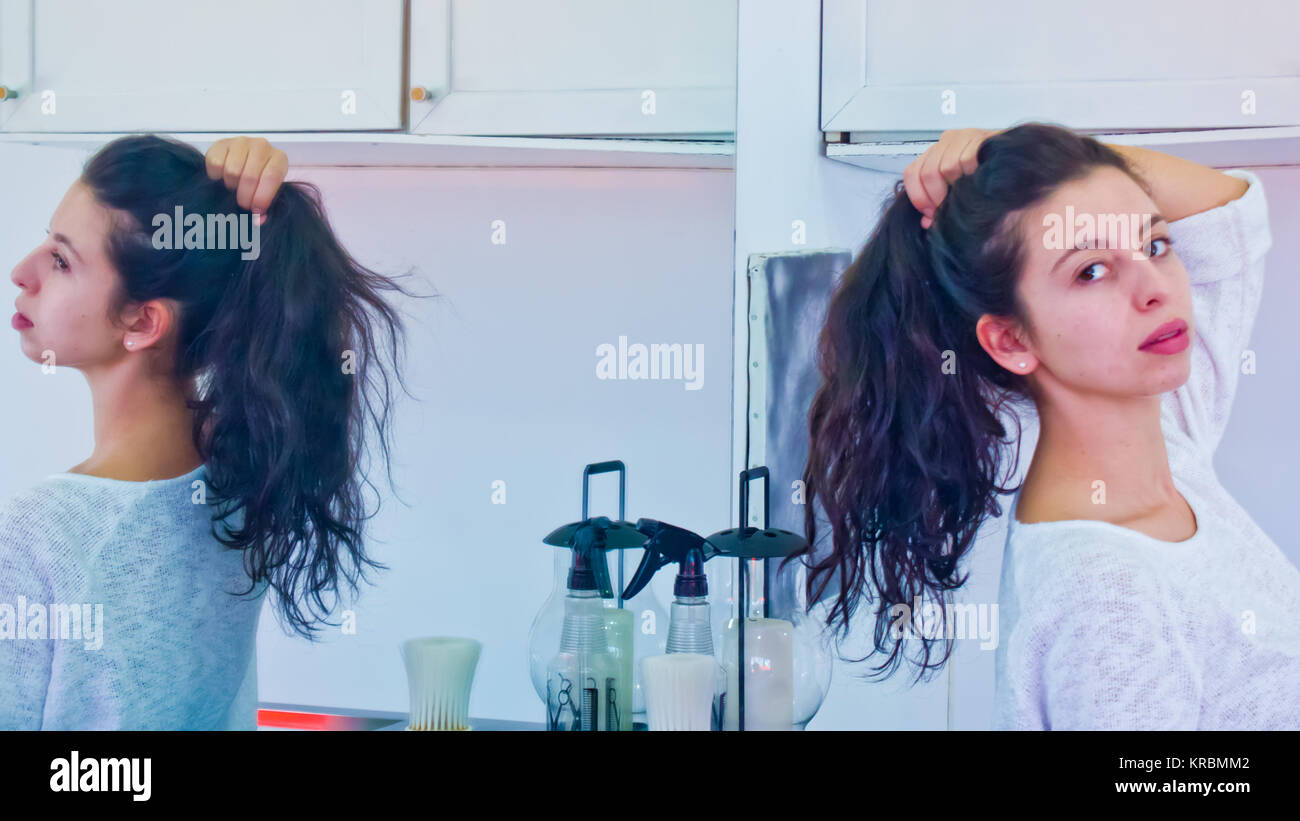 Giovane donna che mostra la sua lunga sventolato capelli scuri riflessa in uno specchio e legati in una coda di cavallo dalla sua propria mano Foto Stock