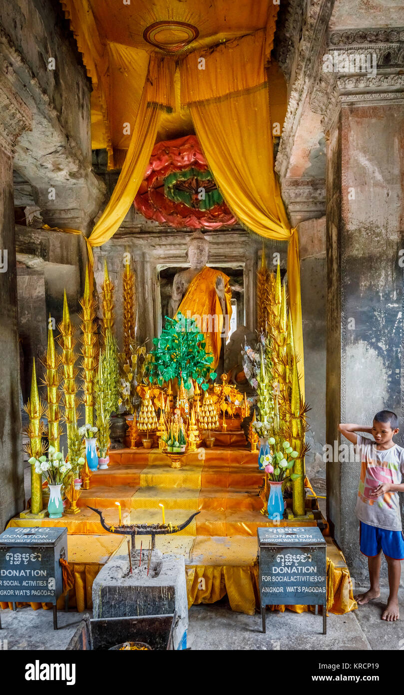 Ragazzo locale in piedi da un colorato santuario buddista con una statua di Buddha all'interno di Angkor Wat pagoda, un tempio complesso vicino a Siem Reap in Cambogia Foto Stock