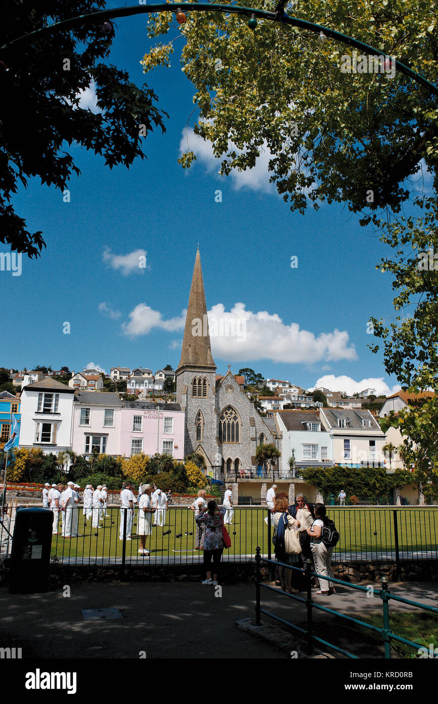 Una partita di bocce si svolge su un verde a Dawlish, nel Devon, dominato da una chiesa con una guglia. I giocatori sono tutti vestiti di bianco tradizionale. Foto Stock