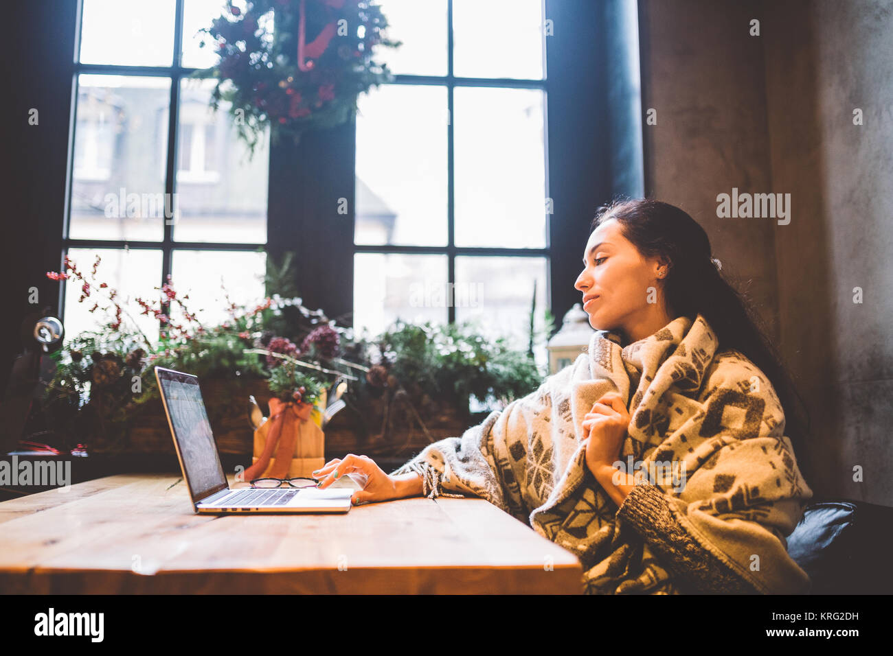 Bella ragazza giovane utilizza la tecnologia portatile, testo tipi guardando il monitor in un caffè dalla finestra al tavolo di legno, in inverno decorata con decorazioni di Natale.vestito con una maglia grigia maglione di lana Foto Stock