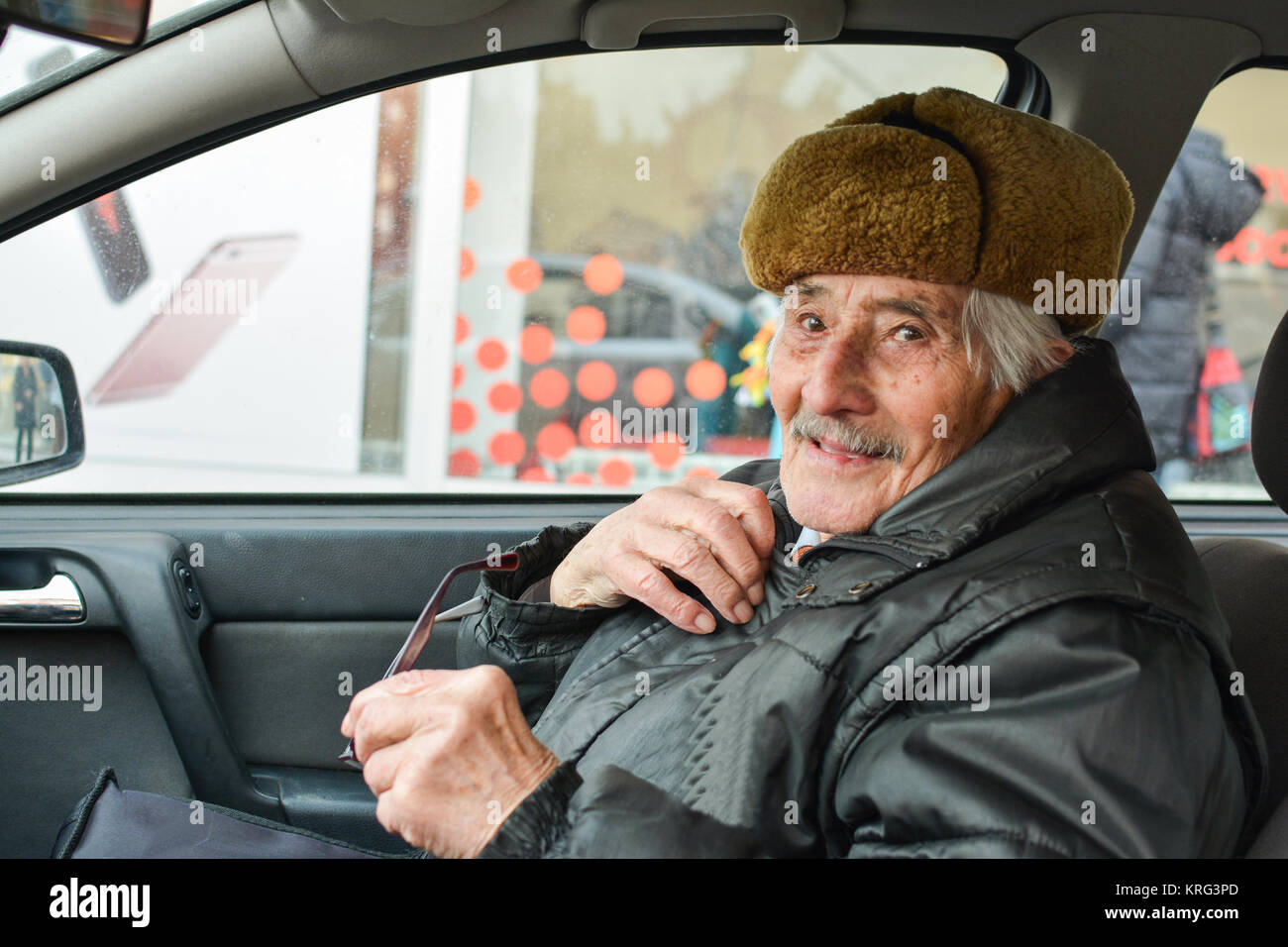 Di vitale importanza uomo anziano con il cappello di pelliccia in un auto,  sorridente e tenendo i bicchieri durante la guida Foto stock - Alamy