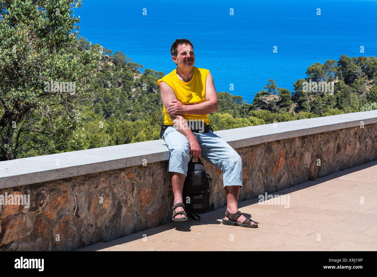 Ein Mann sitzt bei Sonneschein auf einer Mauer im Hintergrund Pinienwald und das blaue Meer. Aufnahme auf Mallorca, Spanien. Foto Stock
