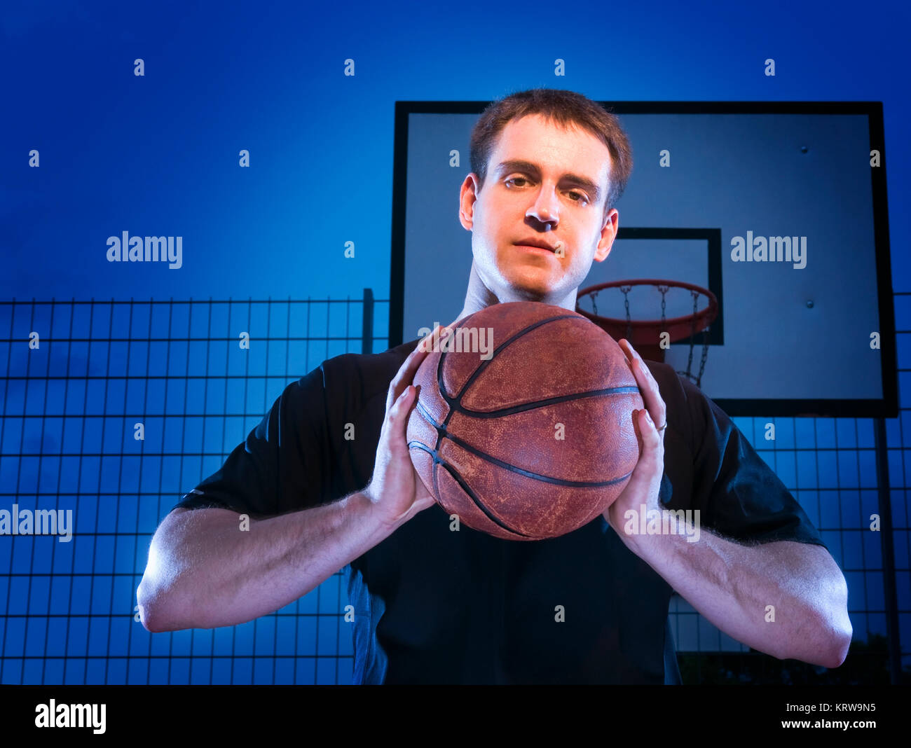 Frontale Oberkörper-Ansicht eines jungen Mannes mit Basket nächtlich vor blauen Hintergrund mit Basketballkorb. Foto Stock