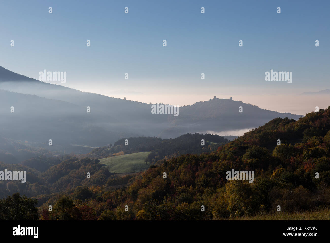 Vista della valle umbra con colline piene di alberi di autunno e la città di Assisi in background, al di sopra di alcuni strati di nebbia Foto Stock