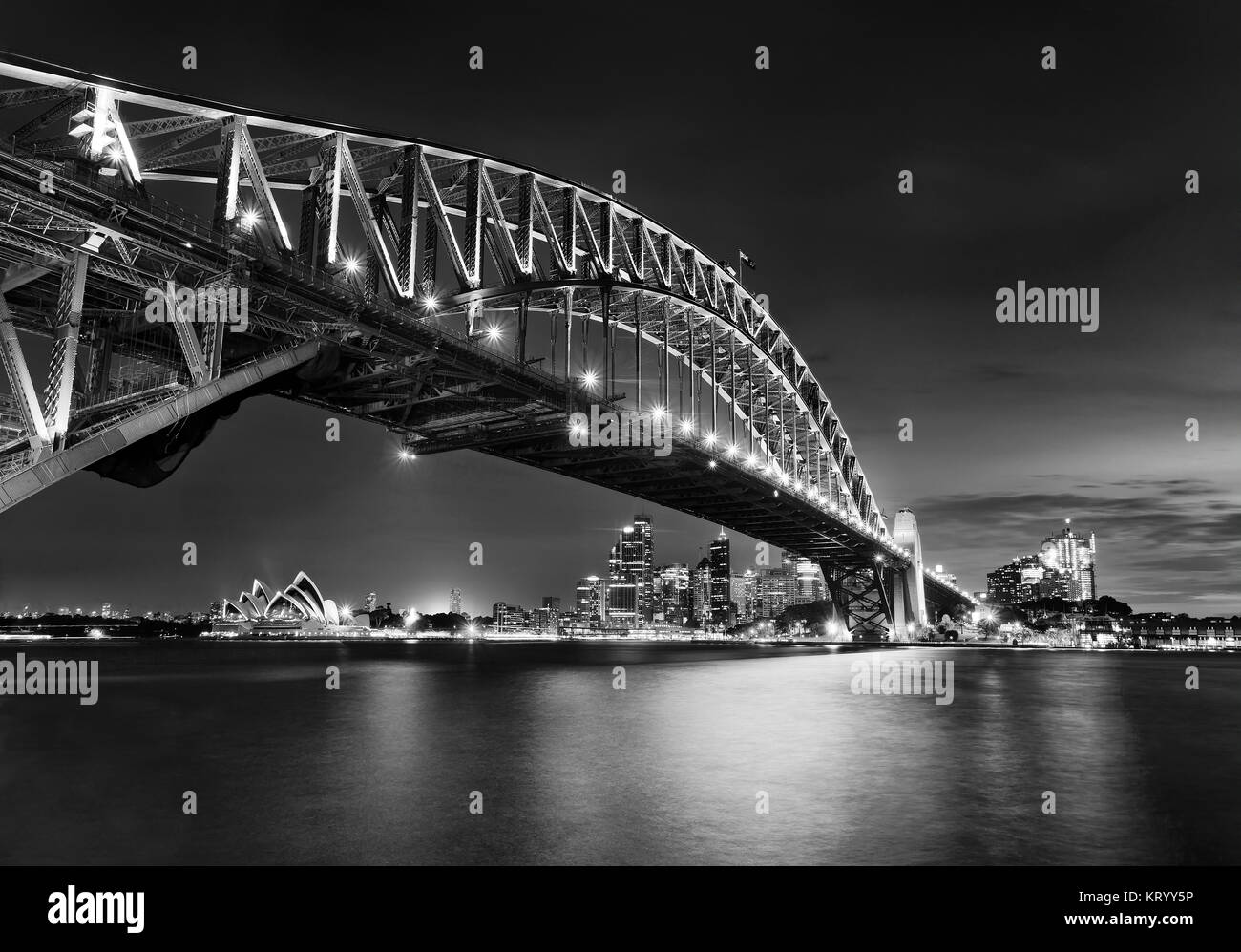 Bianco-nero in vista laterale del Sydney Harbour Bridge verso la città CBD, rocce e Circular Quay al tramonto in tutta sfocata porto riflettente l'acqua. Ponte Foto Stock