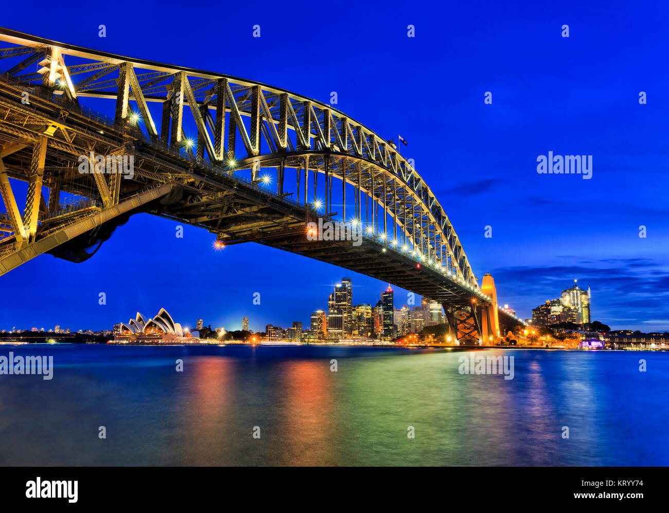 Vista laterale del Sydney Harbour Bridge verso la città CBD, rocce e Circular Quay al tramonto in tutta sfocata porto riflettente l'acqua. Arcata del ponte è malato Foto Stock