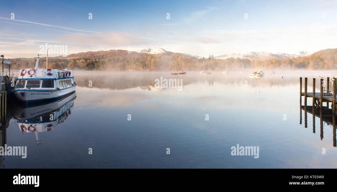 La nebbia si alza dalle tranquille acque del lago di Windermere a Ambleside Pier dove un traghetto barca è ormeggiata, sotto thew montagne innevate di Langdale, E Foto Stock