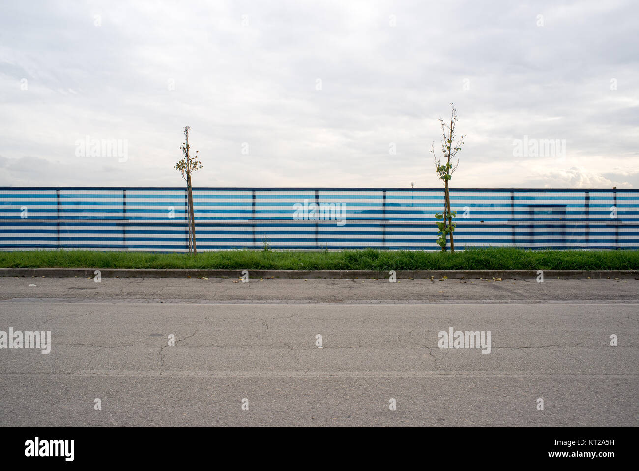 Strada laterale di un blu e bianco striato recinzione su strada con due piccoli alberi con uno sfondo con cielo nuvoloso Foto Stock