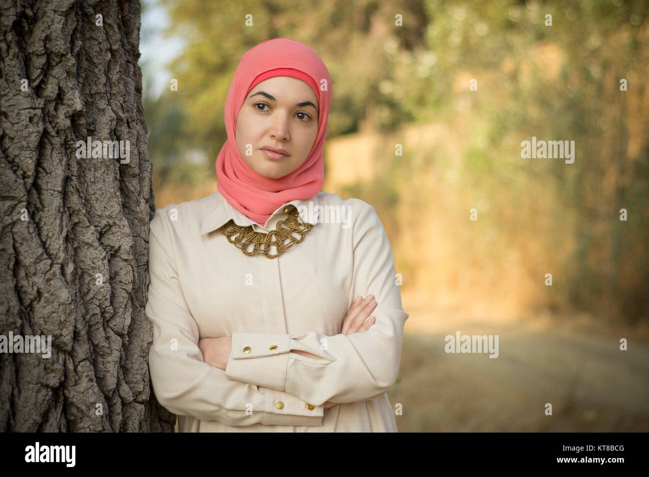 Bella donna musulmana che indossa il hijab appoggiata contro un albero bracci ripiegati che guarda lontano Foto Stock