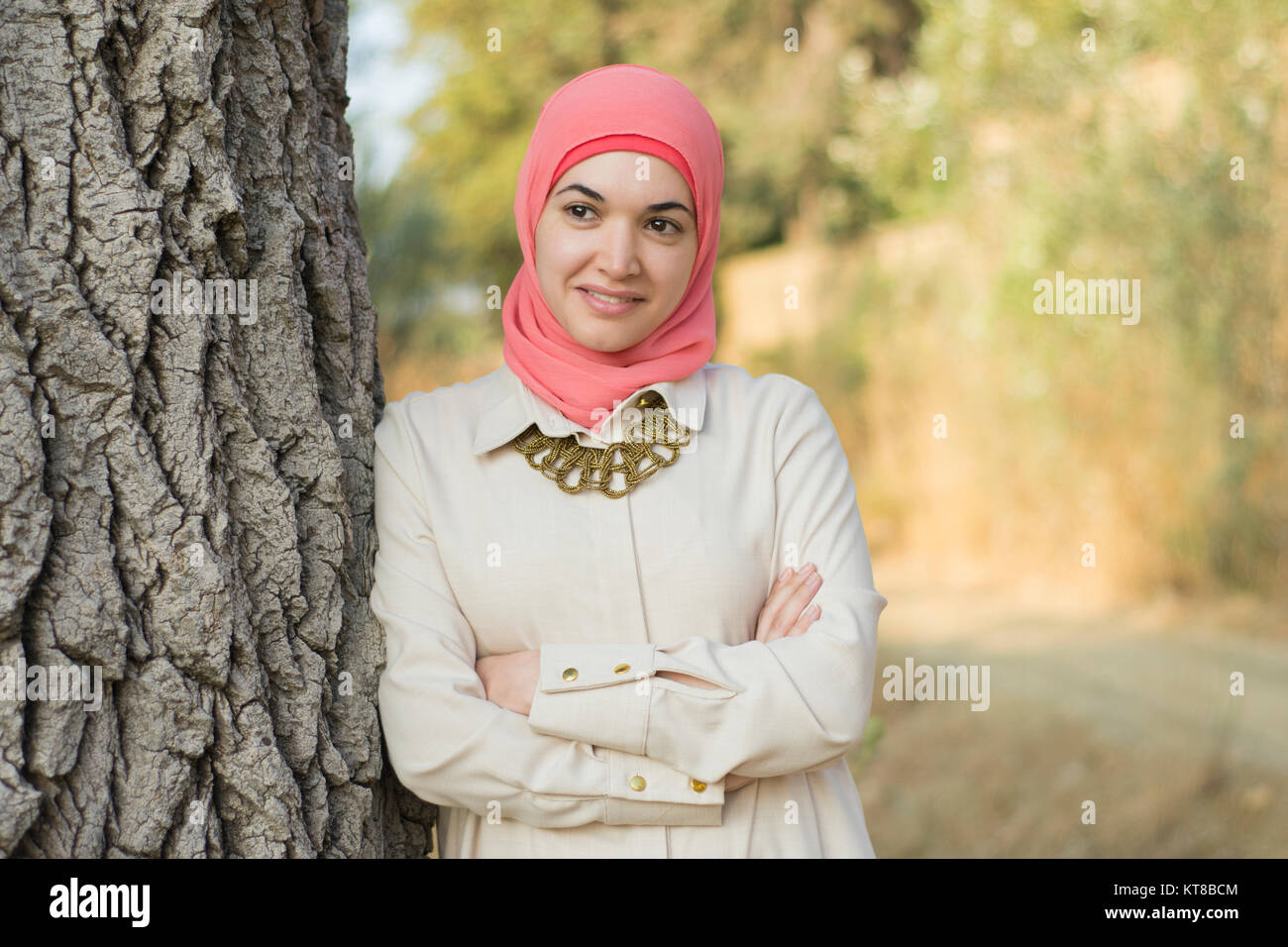 Bella donna musulmana che indossa il hijab appoggiata contro un albero bracci ripiegati che guarda lontano sorridente Foto Stock