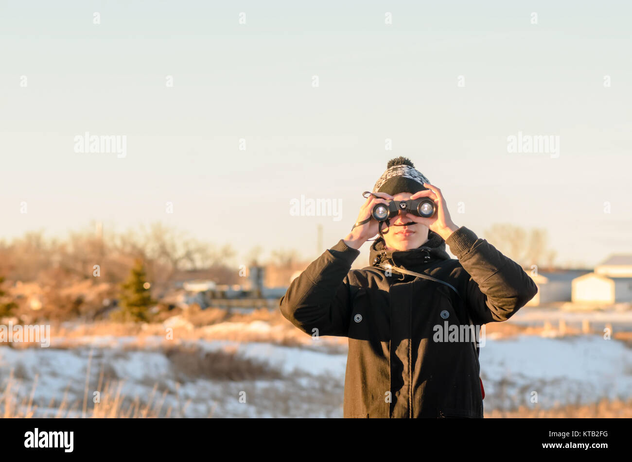 In primo piano, un giovane ragazzo in una giacca nera guarda attraverso il binocolo, sullo sfondo bianco della neve, giallo erba e arbusti, sopra il blu chiaro Foto Stock