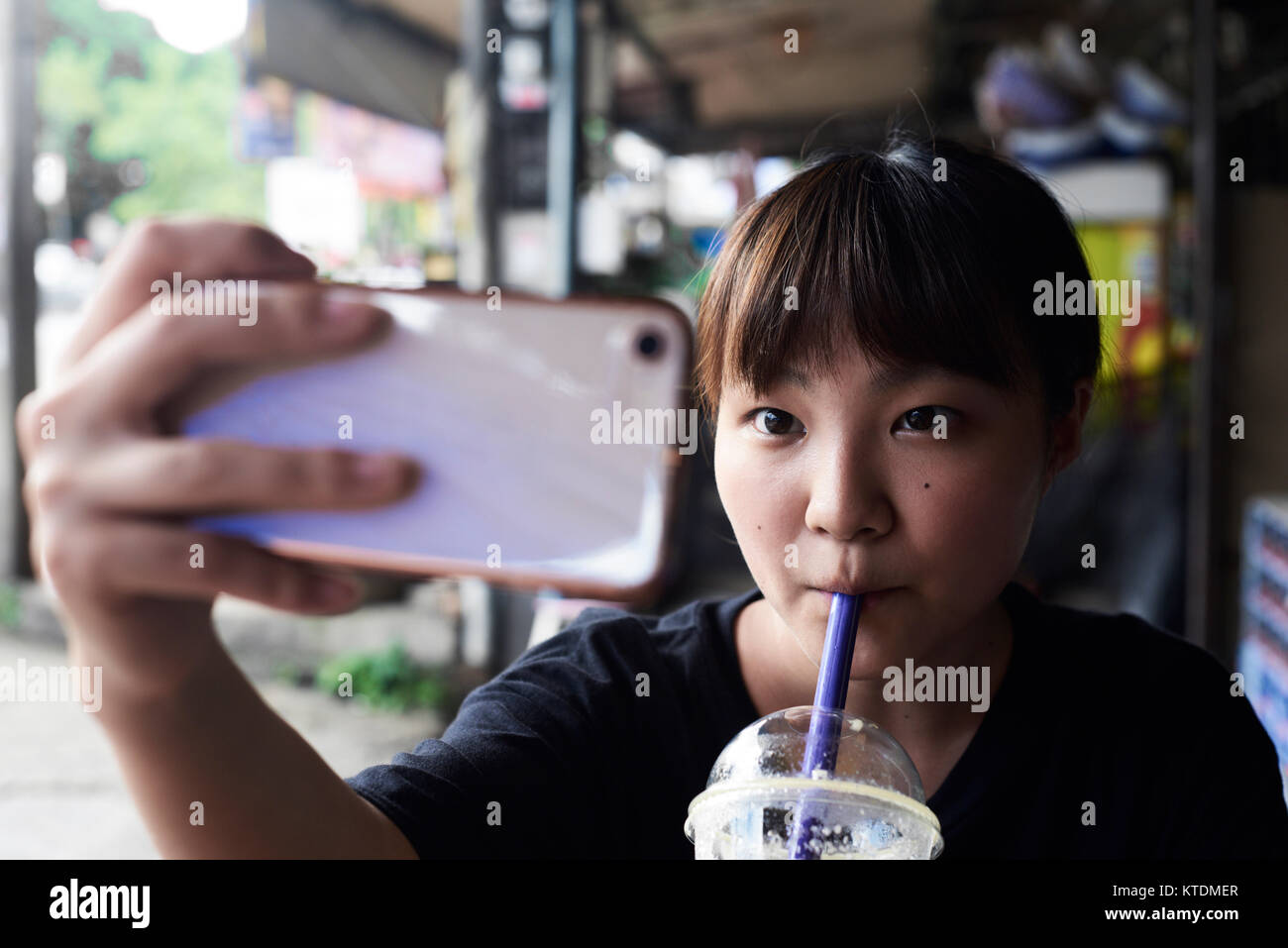 Bella femmina asiatica di bere un smoothie tenendo un autoritratto con il suo telefono cellulare in un ambiente urbano. Chiang Mai, Thailandia. Foto Stock