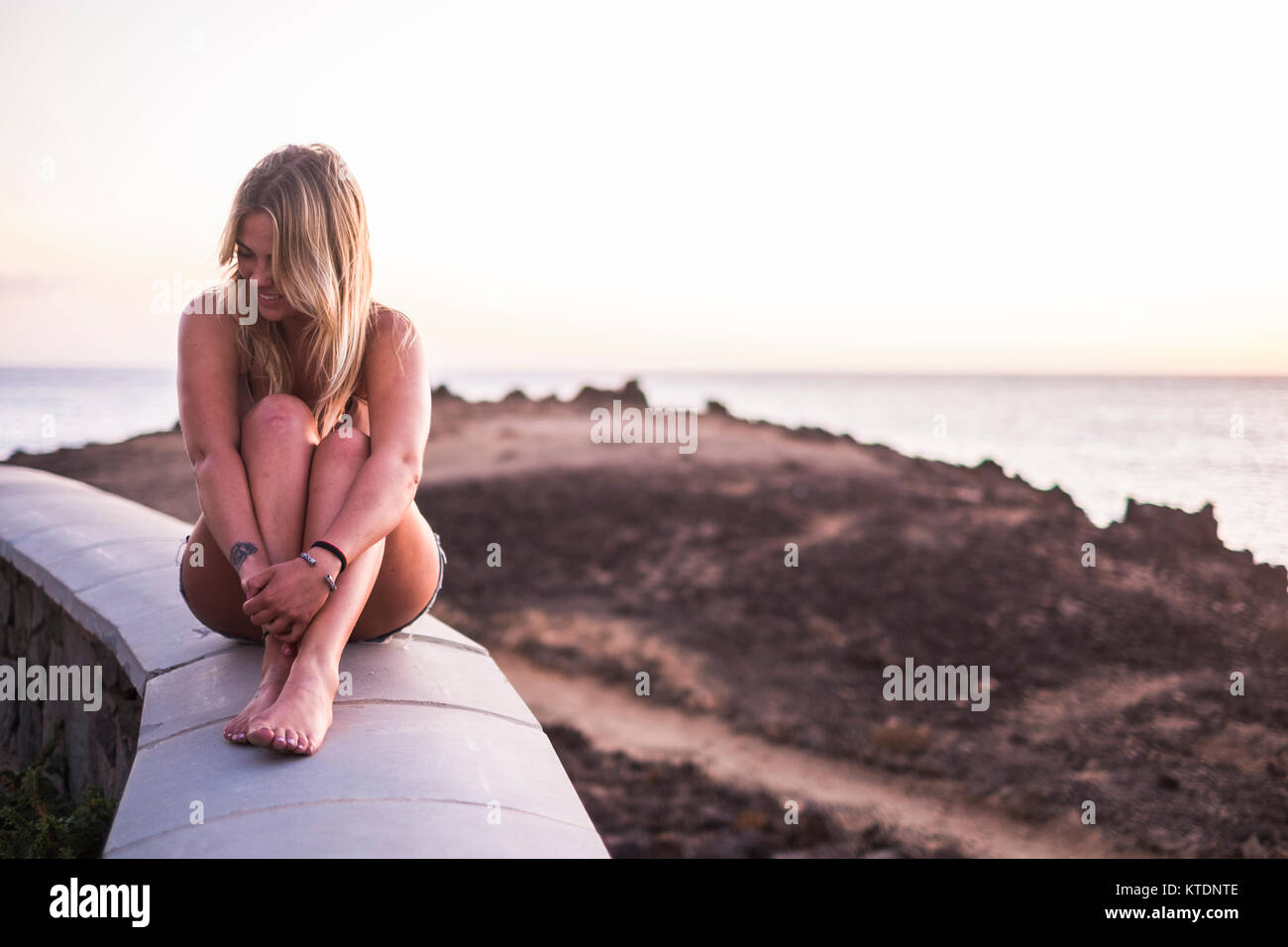 Spagna, Tenerife, ridendo donna bionda seduta su un muro vicino al mare al tramonto Foto Stock