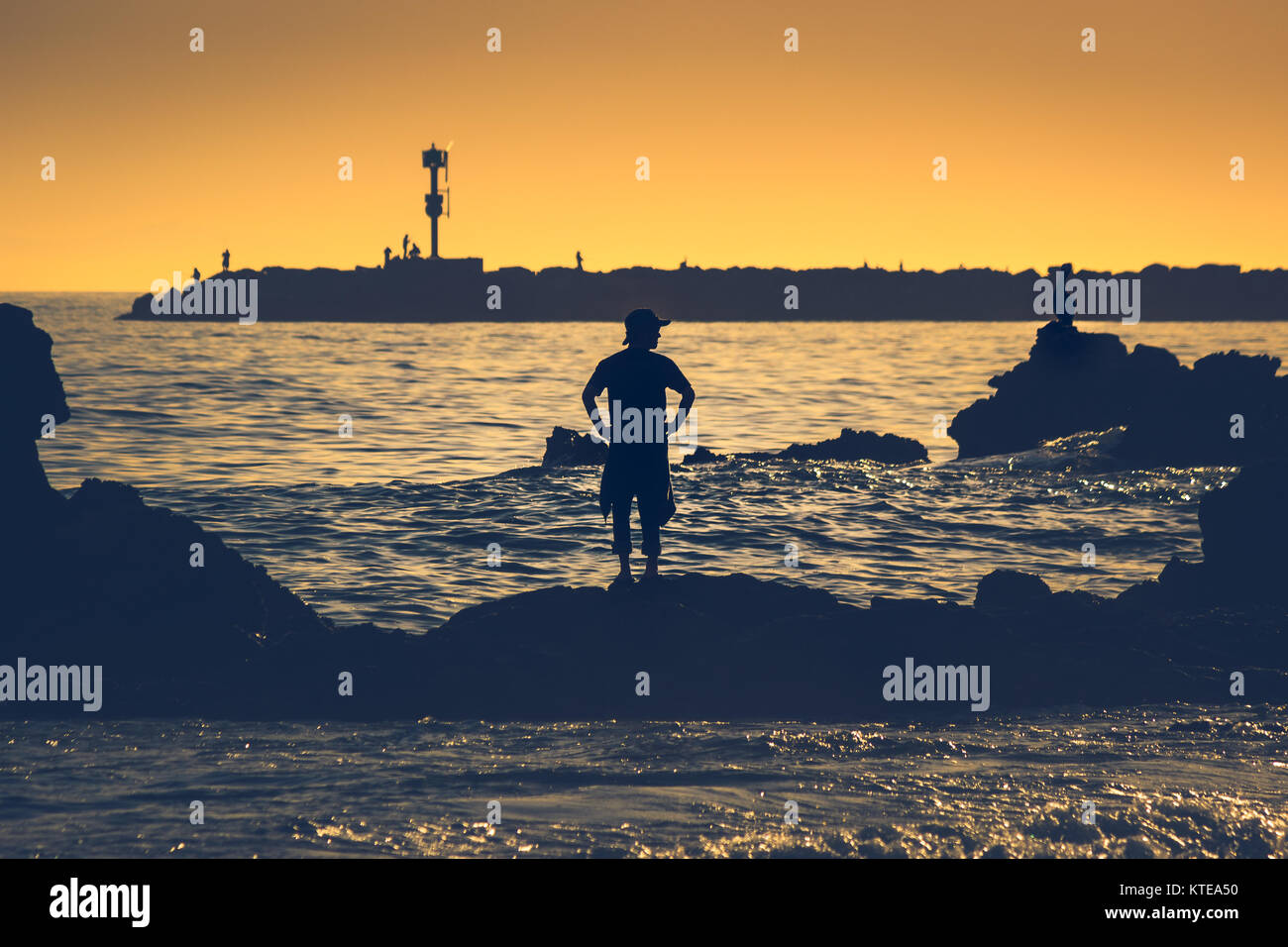 Silhouette di uomo poste su una formazione di roccia al tramonto dalla piccola spiaggia della Corona, Corona del Mar, California con persone di pesca dal molo del bac Foto Stock