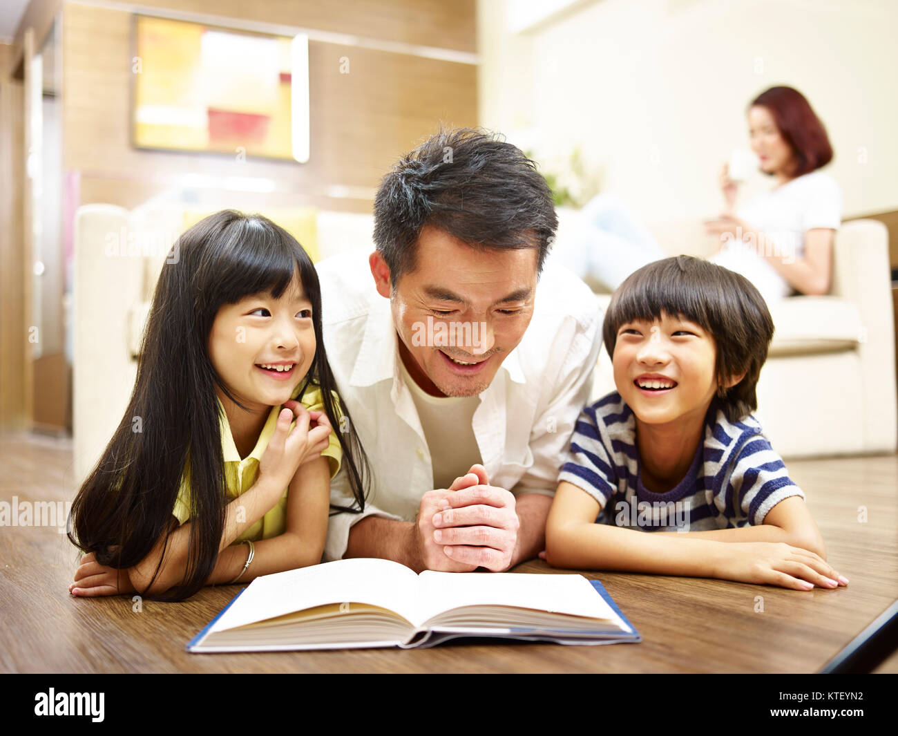 Asian padre e due figli che giace sulla parte anteriore sul pavimento libro lettura mentre madre rilassante in background. Foto Stock