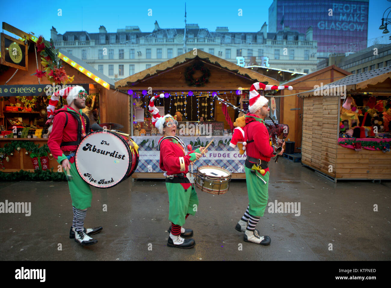 Vento e pioggia salutare i last minute la vigilia di Natale i visitatori per il Natale fayre nella città del George Square.come la commedia pipe band di absurdist marzo attraverso la folla di persone rendere lo sfondo di Glasgow Foto Stock
