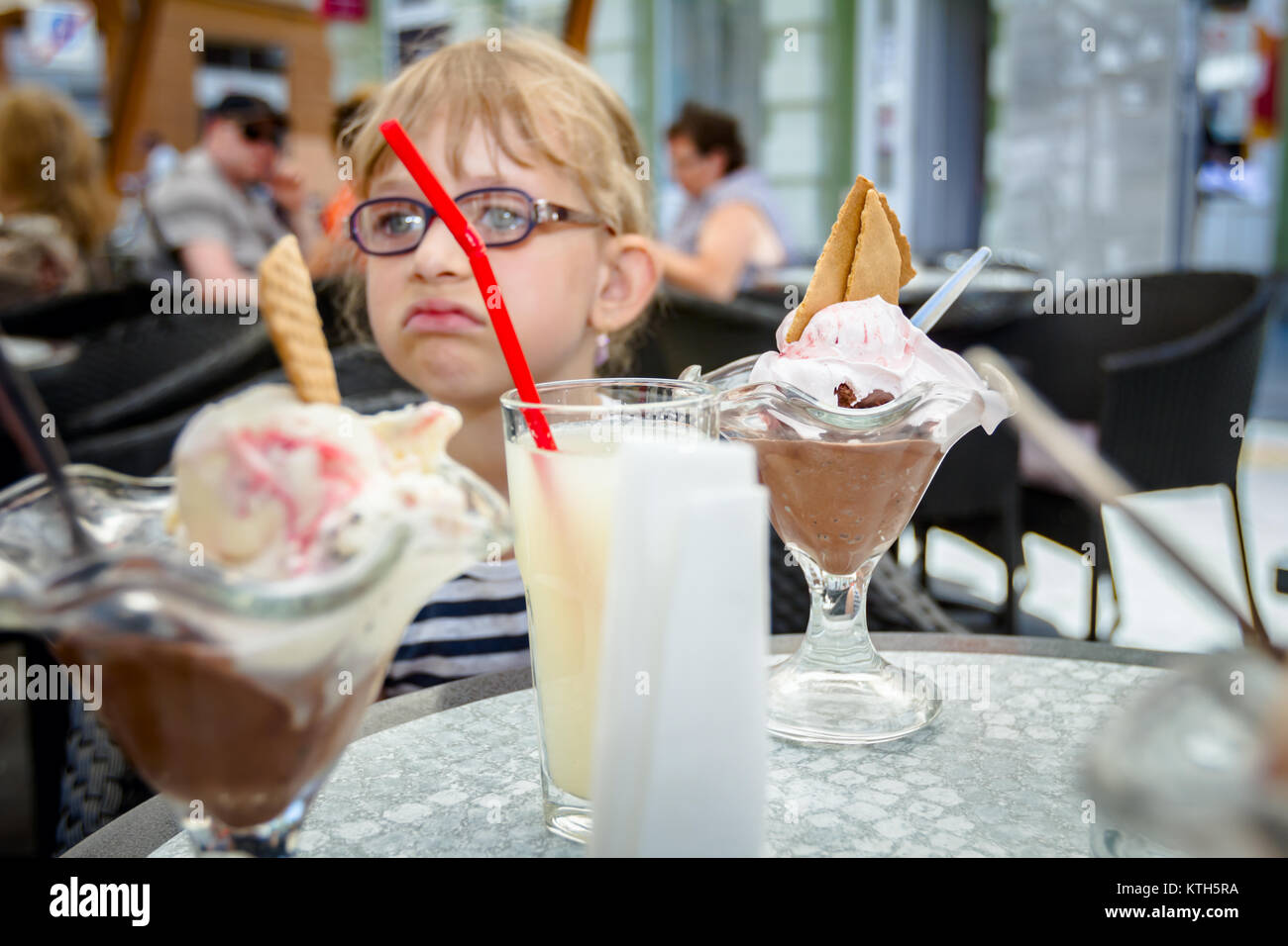 Poco carino ragazza con gli occhiali e la faccia di spensierata espressione è mangiare il gelato al tavolo in una pasticceria in estate. Foto Stock