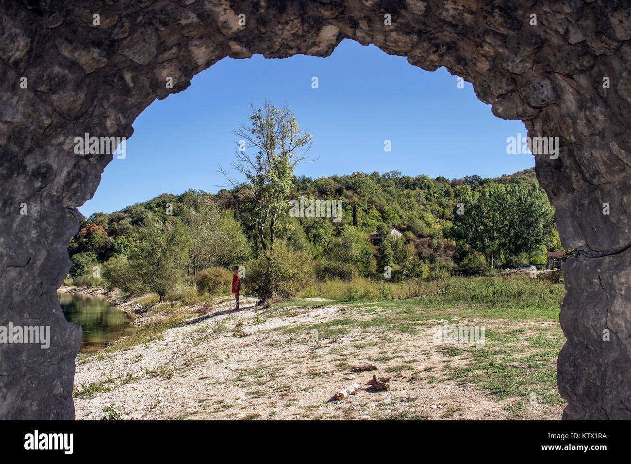 Rijeka Crnojevica, Montenegro - vista attraverso l'arco di un vecchio ponte in pietra di una donna osservando la banca di fiume Foto Stock