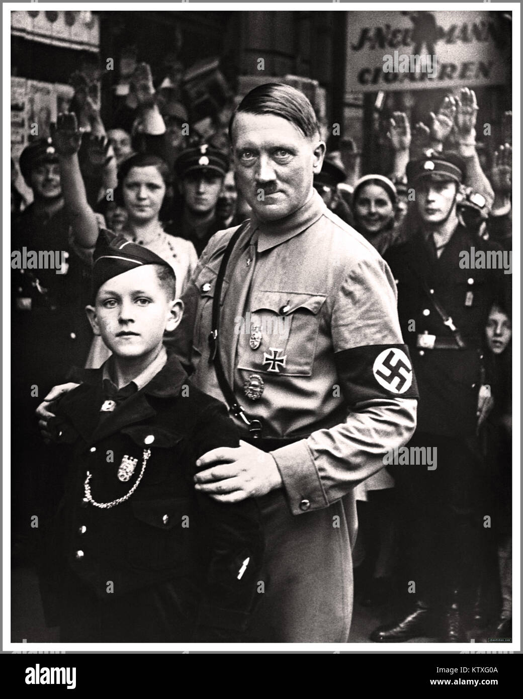 HITLER Germania nazista Propaganda Image 1930 il dittatore nazista Adolf Hitler che indossa l'uniforme NSDAP e la fascia da braccio svastica a Berlino, posando con un'espressione agghiacciante accanto a un giovane membro di 10-12 anni dell'organizzazione Hitler Youth Hitler-Jugend. La folla che saluta Heil Hitler. Foto Stock