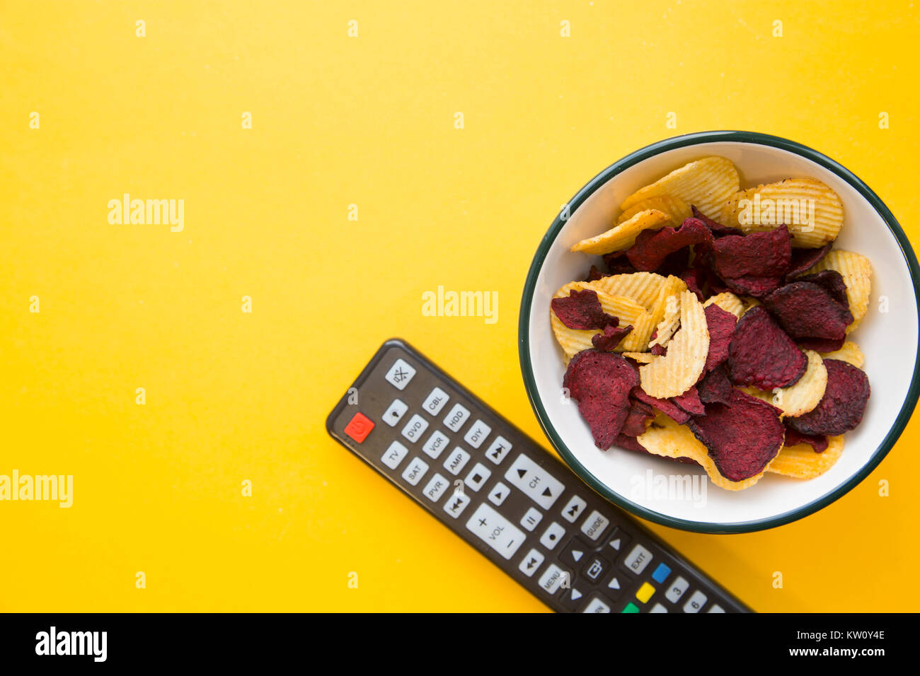 Weekend, hobby e tempo libero concetto. Un colore di sfondo giallo con una ciotola di patate e barbabietole in chip e un telecomando del televisore. Spazio per il testo Foto Stock Weekend, hobby e tempo libero concetto. Un colore di sfondo giallo con una ciotola di patate e barbabietole in chip e un telecomando del televisore. Spazio per il testo Foto Stock