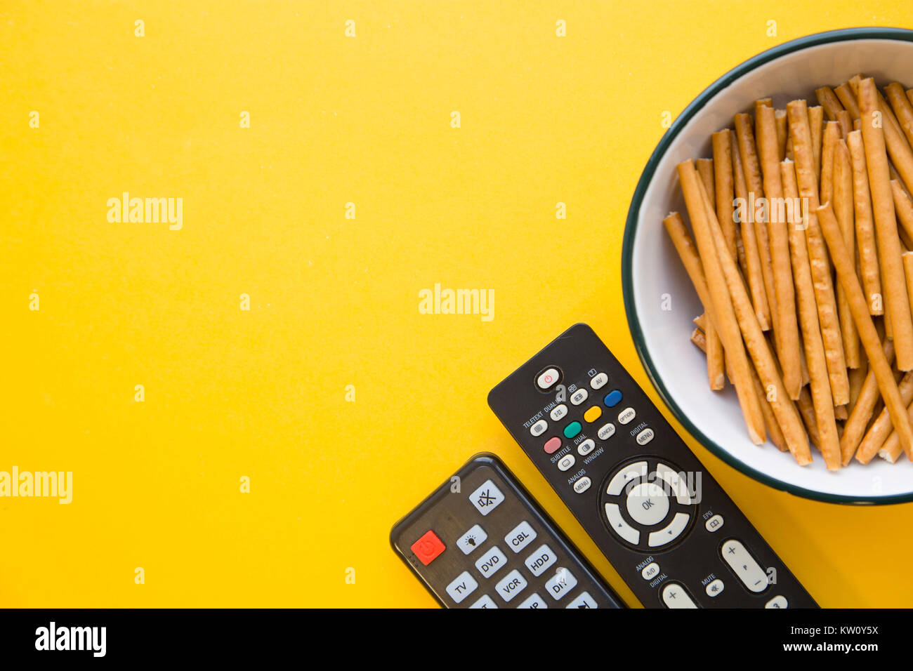 Weekend, hobby e tempo libero concetto. Un colore di sfondo giallo con una ciotola di pane salato bastoni e due telecomandi della TV. Lo spazio per il tuo testo o Foto Stock