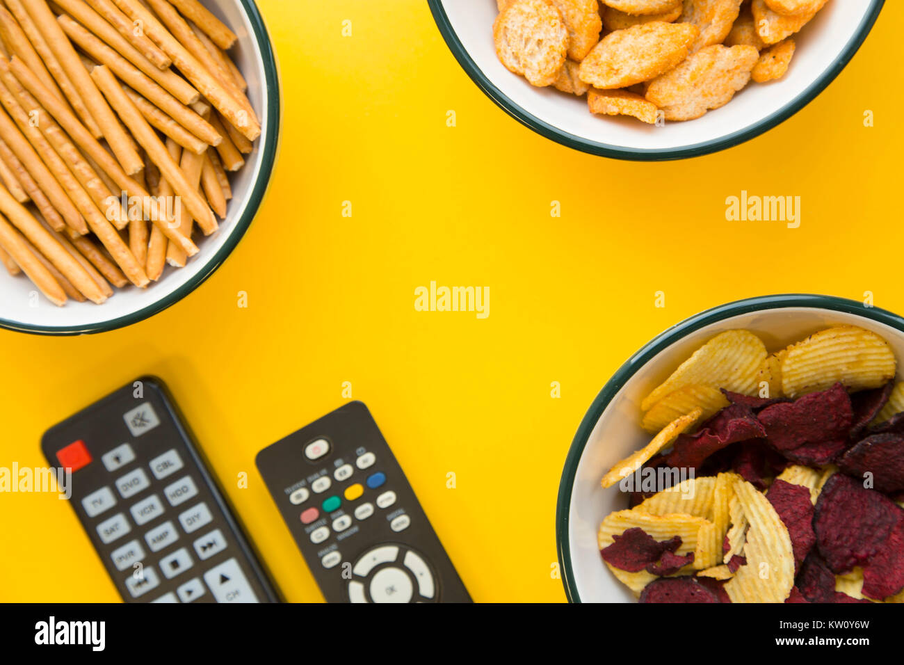 Una serata con gli amici davanti alla tv. Tre ciotole con pane salato di bastoni, briciole di pane e patatine e due telecomandi su un giallo brillante bac Foto Stock