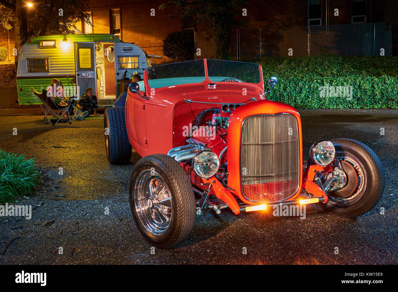 1932 red Ford hot rod roadster di notte con rimorchio camper e persone rilassante in background durante la notte. Foto Stock