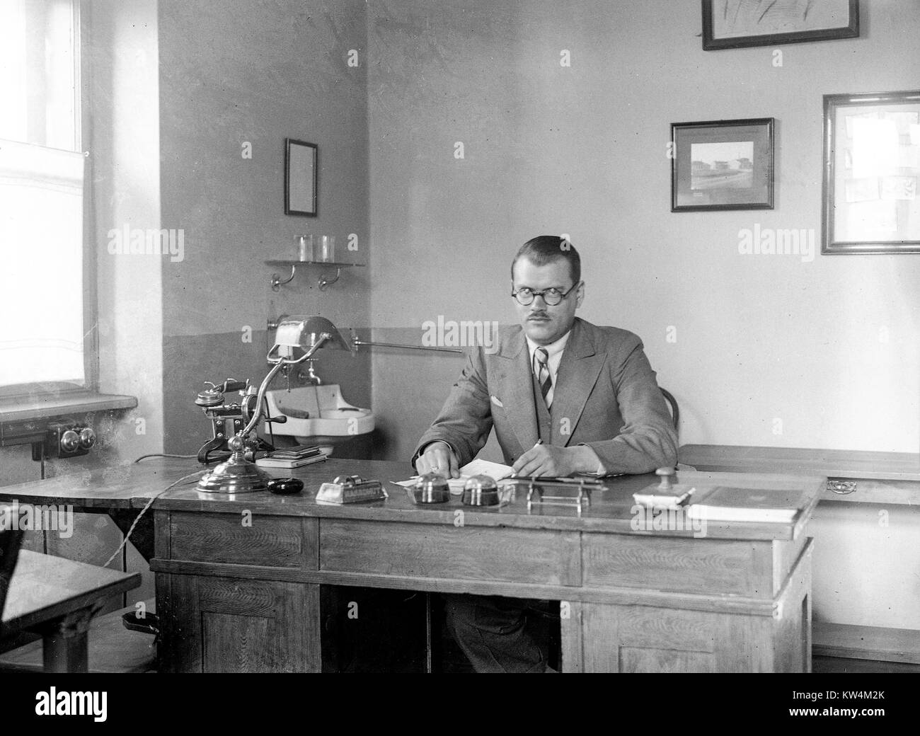 Un uomo con capelli corti, round occhiali una tuta conservatore e a poppa con un espressione del viso si siede dietro una grande scrivania di legno nel suo ufficio, con un piccolo lavabo, un inchiostro blotter, due campane, una scrivania, una lampada e un wind-up telefono visibile, Ungheria, 1929. Digitalizzate da un processo di gelatina lastra di vetro negativo. Foto Stock