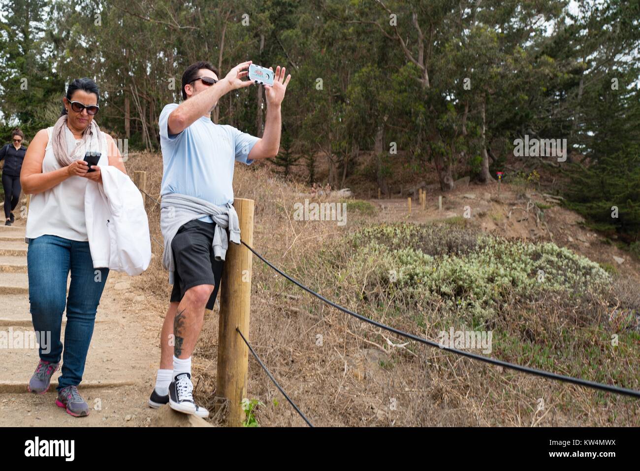 Voce maschile turistica prendendo un cellulare foto del Golden Gate mentre una femmina passeggiate turistiche passato e utilizza il suo telefono cellulare, il sentiero costiero di Lands End quartiere di San Francisco, California, 27 agosto 2016. Foto Stock