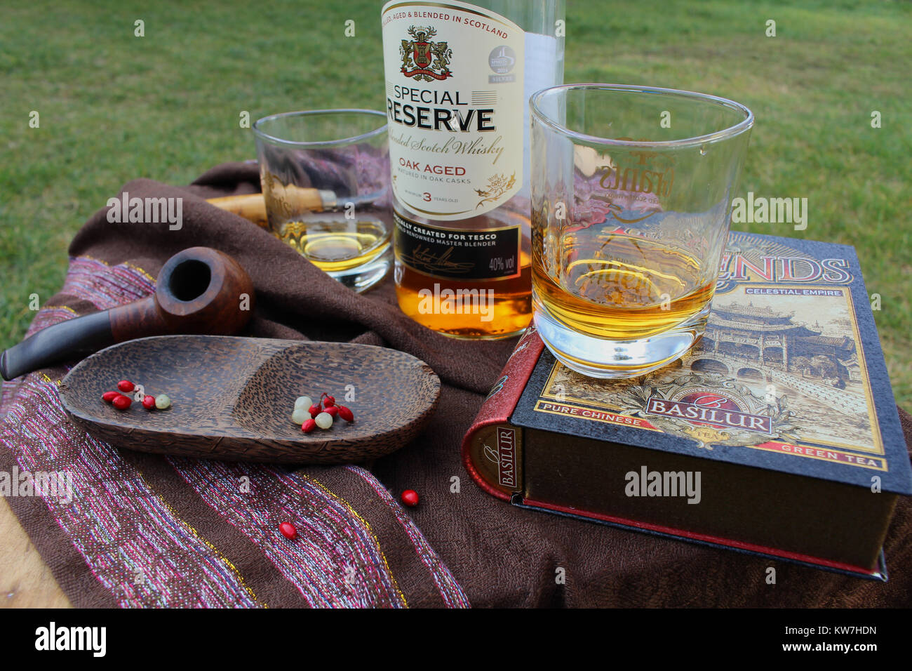 Still Life - due bicchieri con bottiglia di whisky, tubo di legno e ciotola e un libro disposti su un tavolo da giardino Foto Stock