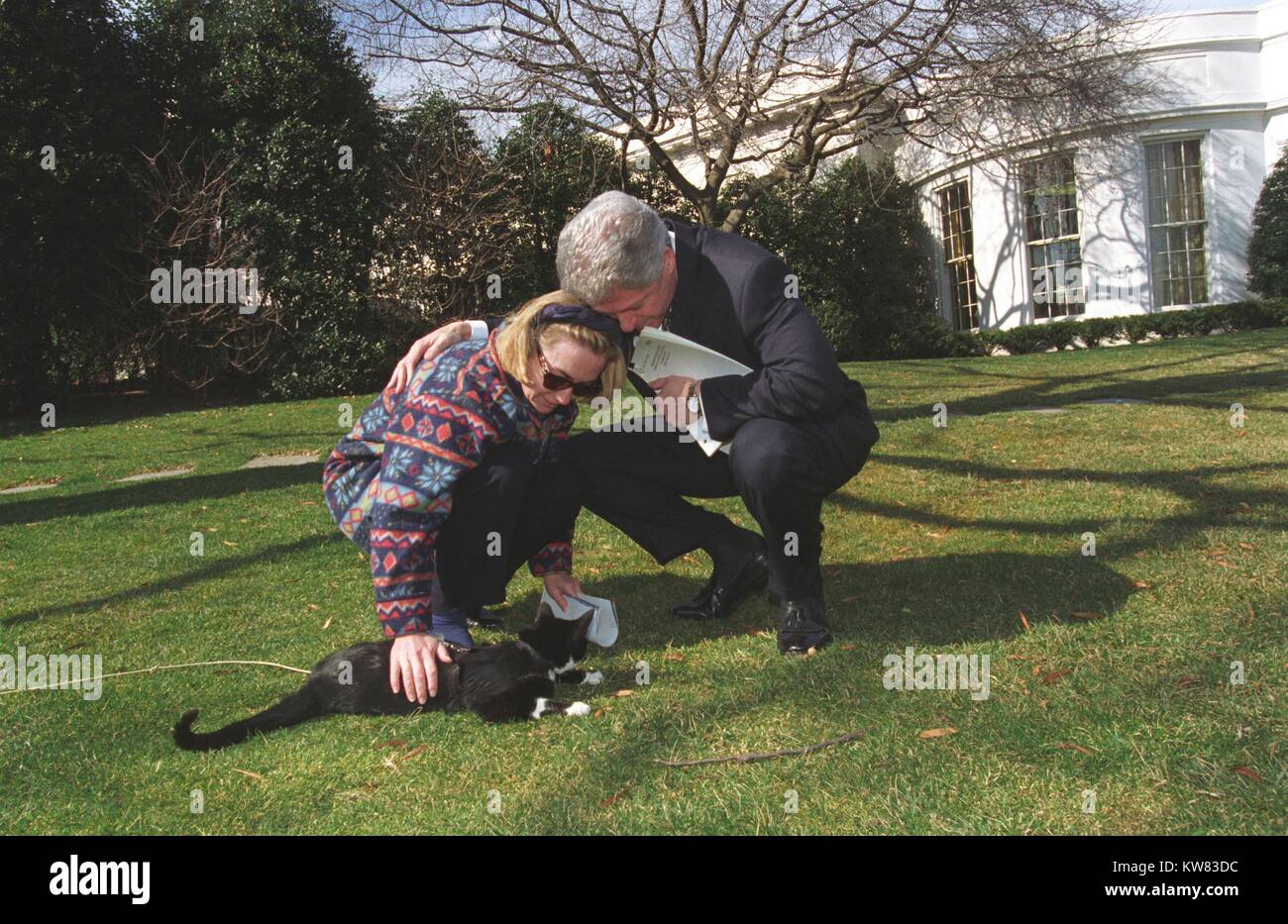 Sul prato davanti alla Casa Bianca il Presidente Bill Clinton indossando abito nero e tenendo la carta si piega verso il basso a fianco della prima signora Hillary Clinton modellato da indossare pullover e occhiali da sole mentre lei lo accarezza il loro primo Pet, calze il gatto con pelo nero, faccia bianca, e string guinzaglio serpeggiava in erba, febbraio 24, 1997. Foto Stock