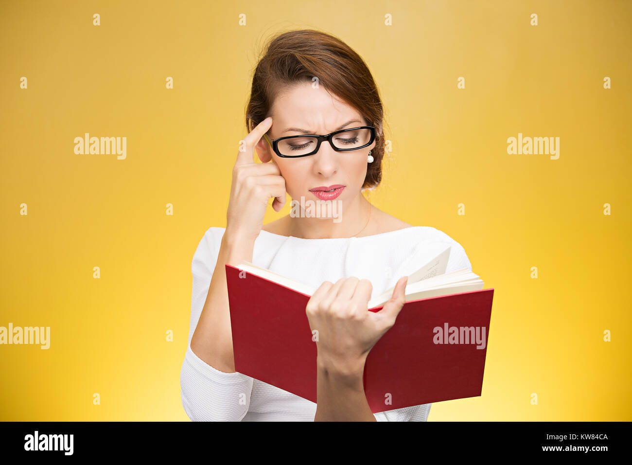 Giovane donna elegante in occhiali aventi problemi con la comprensione durante la lettura del libro isolato su sfondo giallo Foto Stock