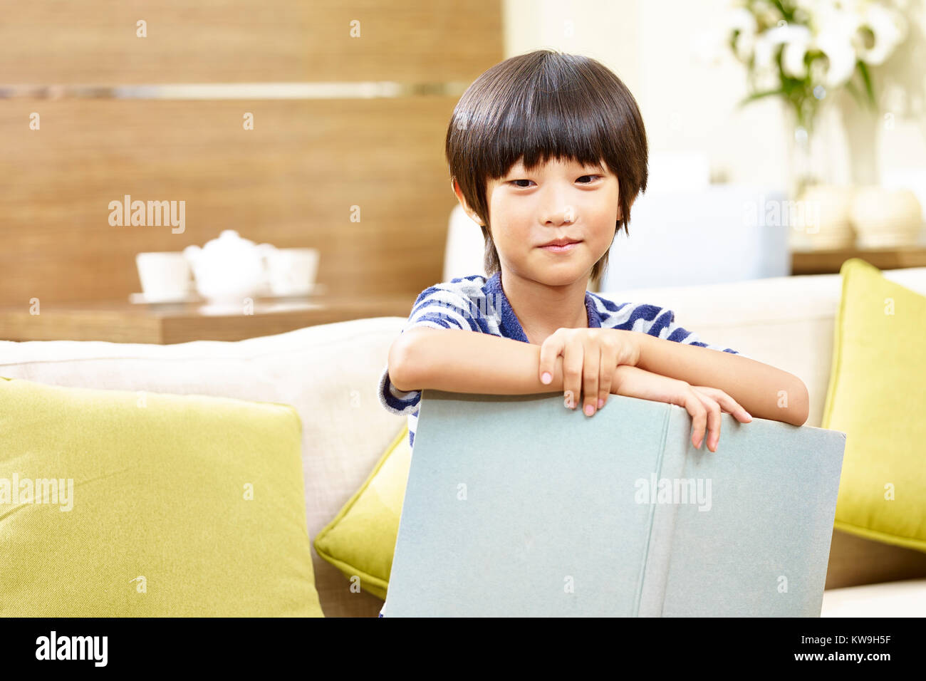 8-anno-vecchio piccolo ragazzo asiatico seduto sul divano con un libro in mano guardando la telecamera sorridendo. Foto Stock