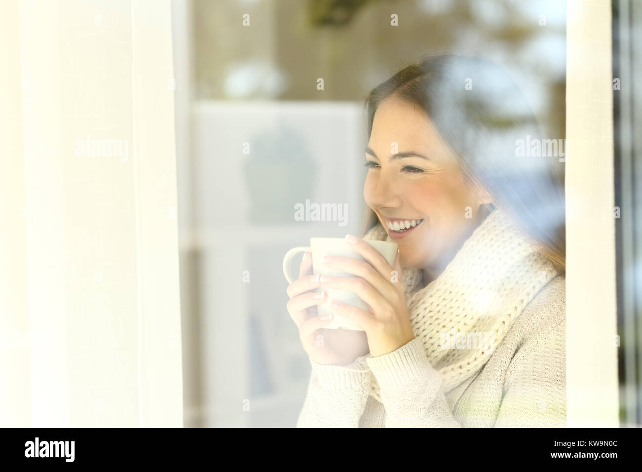Donna felice di guardare attraverso una finestra tenendo una tazza da caffè in piedi a casa in inverno Foto Stock