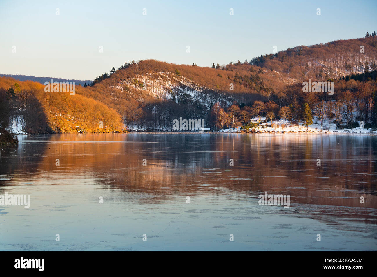 Vista serale oltre l'Obersee congelata del lago Rursee con la riflessione del paesaggio illuminato in inverno in Eifel, Germania. Foto Stock