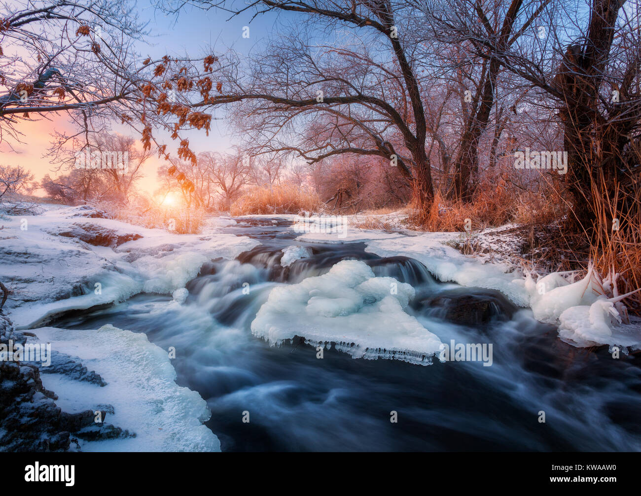Foresta di inverno con il maestoso fiume al tramonto. Paesaggio invernale con alberi innevati, ghiaccio, bellissimo fiume congelato, boccole nevoso e colorati di cielo nel crepuscolo. Blurre Foto Stock