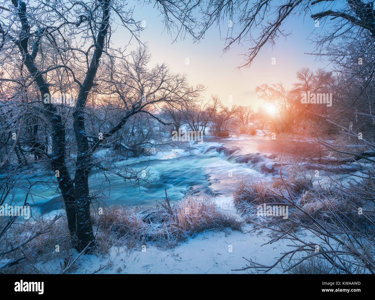 Foresta di inverno con il maestoso fiume al tramonto. Paesaggio invernale con alberi innevati, ghiaccio, bellissimo fiume congelato, boccole nevoso e colorati di cielo nel crepuscolo. Blurre Foto Stock