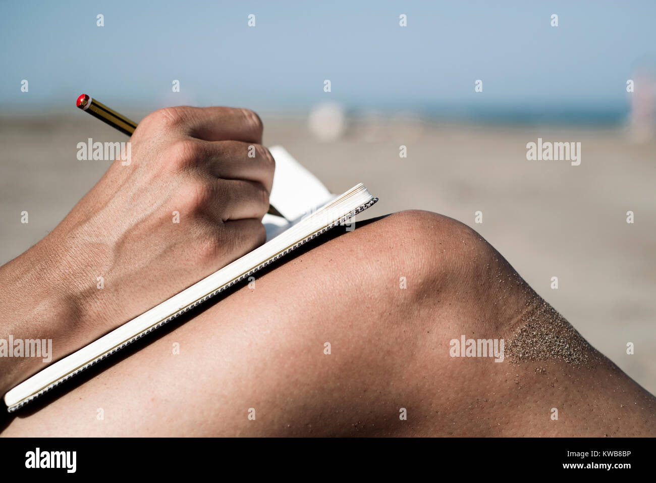 Primo piano di un giovane uomo caucasico scrivere con una penna in un notebook sulle sue gambe rasata, sulla spiaggia durante la stagione estiva Foto Stock