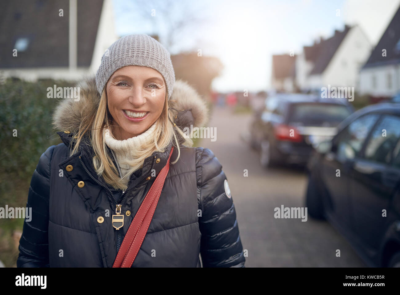 Sorridendo felice di mezza età donna in piedi all'aperto in una fredda giornata invernale in una giacca calda e cappuccio a maglia in una strada suburbana Foto Stock