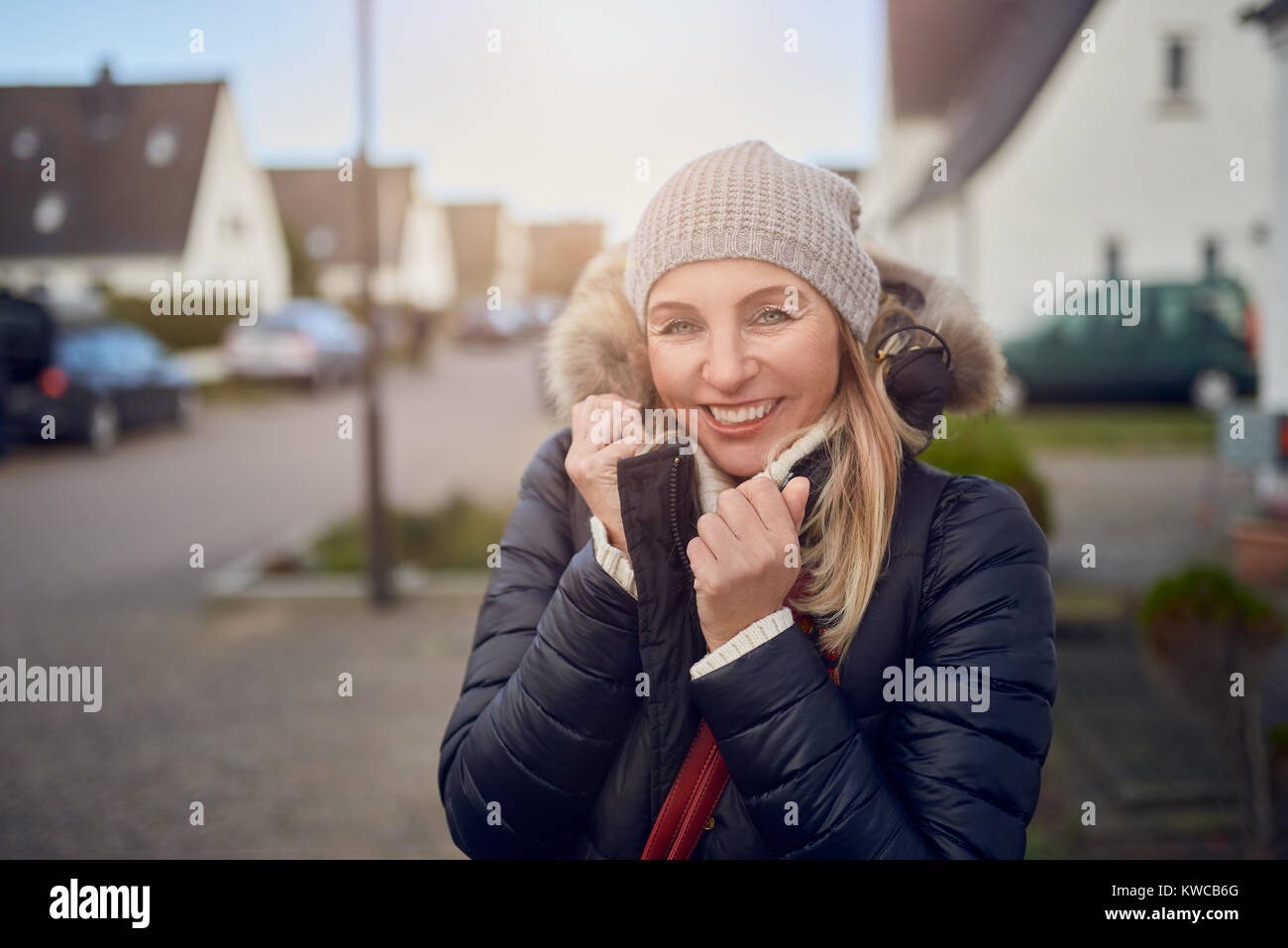 Sorridendo felice di mezza età donna in piedi all'aperto in una fredda giornata invernale in una giacca calda e cappuccio a maglia in una strada suburbana Foto Stock